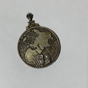 May include: A silver-toned pendant featuring a detailed depiction of a globe, with continents and gridlines. The pendant includes a loop for hanging and a design of flags. The overall design is circular.