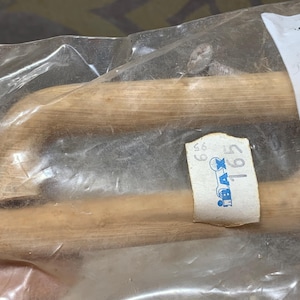 Pode incluir: Duas puxadores de armário de madeira castanho claro são visíveis através de um saco plástico transparente. As puxadores têm um design curvo. Uma etiqueta de preço com o texto "BAX 165" está anexada.