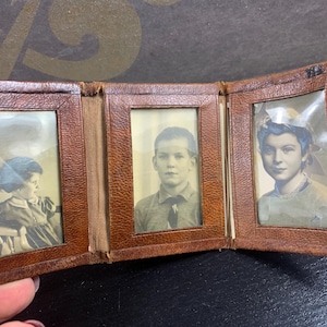 May include: A vintage brown leather photo album, open to display three black and white portraits. The album has a textured surface and a worn appearance, suggesting age. The portraits feature individuals in classic attire.