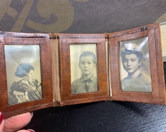 Puede incluir: Un álbum de fotos vintage de cuero marrón, abierto para mostrar tres retratos en blanco y negro. El álbum tiene una superficie texturizada y un aspecto desgastado, lo que sugiere antigüedad. Los retratos muestran personas con vestimenta clásica.
