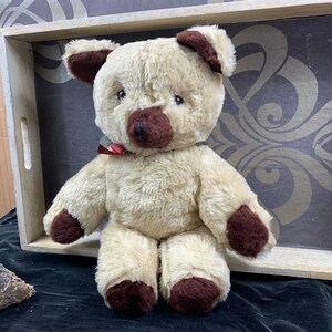 May include: A beige teddy bear with brown ears and paws, sitting on a wooden tray with a patterned background.