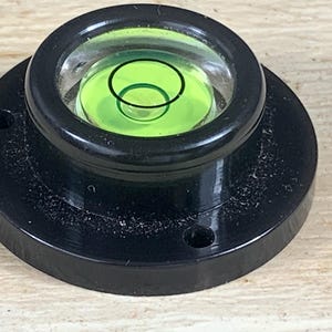 Puede incluir: Un nivel de burbuja circular negro con una parte superior transparente en forma de cúpula. El nivel contiene un líquido verde brillante y un indicador circular negro. La base tiene dos orificios de montaje. La herramienta es para medir superficies niveladas.