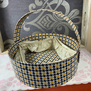 Op de afbeelding: Een vintage hoedenbox met een zwart-geel ruitpatroon en een handvat. De doos is gevoerd met stof en is verdeeld in compartimenten.