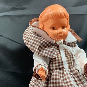 May include: A vintage doll with a brown and white gingham dress and a white lace collar. The doll has a light brown complexion and brown eyes.