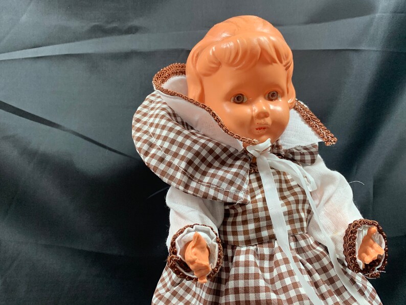 May include: A vintage doll with a brown and white gingham dress and a white lace collar. The doll has a light brown complexion and brown eyes.