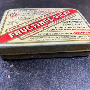 May include: A vintage tin box for Fructines-Vichy laxative candies. The box is yellow with red and gold accents. The text on the box reads "Traitement de la Constipation et de ses consequences" and "Laxative Aromatise aux Sucs de Fruits Agreables a Prendre".