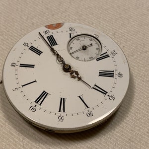 Può includere: Un quadrante di orologio da tasca bianco con indici orari romani neri e indici dei minuti neri. L'orologio ha un piccolo quadrante ausiliario bianco con lancette nere. Il quadrante dell'orologio è leggermente danneggiato.
