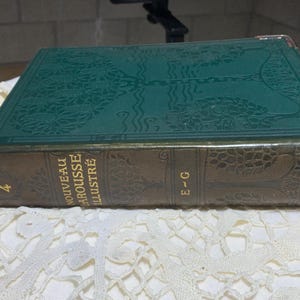 Op de afbeelding: Een vintage hardcover boek met een groene kaft en bruine rug. De kaft heeft een reliëf ontwerp. Op de rug staat de tekst "NOUVEAU LAROUSSE ILLUSTRE" en de letters "E-G". Het nummer "4" is zichtbaar.