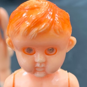 Pode incluir: Close-up do rosto de uma boneca de pl&aacute;stico com cabelo laranja e olhos castanhos. A boneca tem um sorriso pintado e bochechas rosadas.