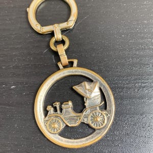 Könnte beinhalten: Vintage Schlüsselanhänger aus Messing mit einem detaillierten Relief eines Oldtimers in einem kreisförmigen Rahmen. Der Schlüsselanhänger enthält einen Verschluss und einen Verbindungsring. Das Auto hat Speichenräder und ein überdachtes Dach. Das Metall hat ein leicht gealtertes Aussehen.