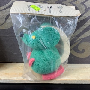 Peut inclure: Un jouet éponge en forme de souris verte avec des pieds roses et un œil jaune, emballé dans un sac en plastique transparent. Le jouet a une poignée circulaire blanche. L'emballage comporte des illustrations d'outils de nettoyage.
