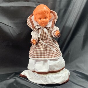 May include: A vintage plastic doll wearing a brown and white gingham dress with a white lace trim and a brown ribbon around the waist.
