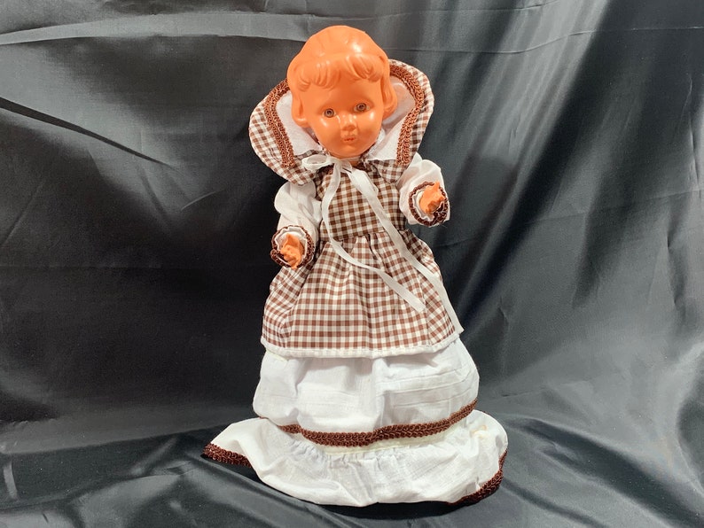 May include: A vintage plastic doll wearing a brown and white gingham dress with a white lace trim and a brown ribbon around the waist.