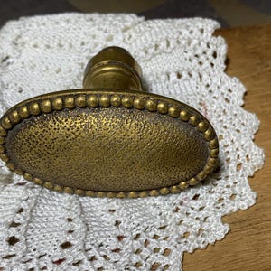 May include: An antique brass door knob with an oval, textured handle and a beaded border. The knob has a tarnished, aged appearance, suggesting it is vintage or a reproduction. The knob is likely for a cabinet or drawer.