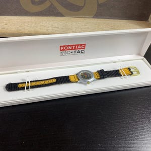 Peut inclure: Une montre Pontiac Tic-Tac dans un coffret blanc. La montre a un bracelet noir et jaune et un cadran argenté. Le coffret porte le logo Pontiac Tic-Tac en rouge et blanc.