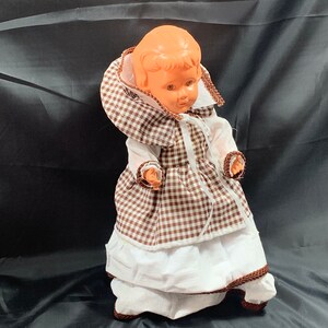 May include: A vintage plastic doll wearing a brown and white gingham dress with a white lace trim. The doll has a brown and white gingham bonnet and a white lace collar.