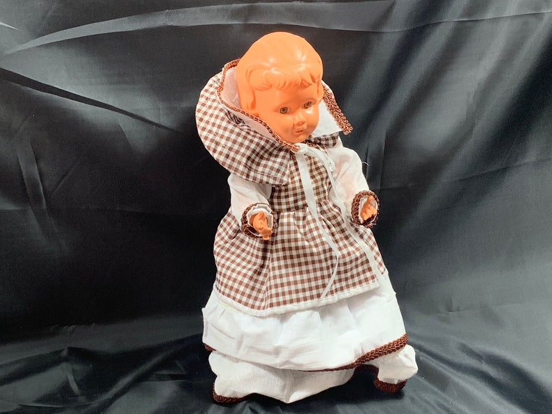 May include: A vintage plastic doll wearing a brown and white gingham dress with a white lace trim. The doll has a brown and white gingham bonnet and a white lace collar.