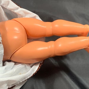 May include: Close-up of a doll's legs, showing the plastic construction and the smooth, peach-colored skin tone. The doll is wearing a white dress with brown and white gingham trim.