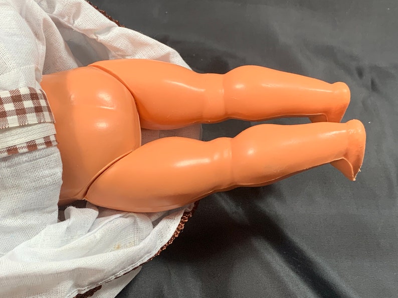 May include: Close-up of a doll's legs, showing the plastic construction and the smooth, peach-colored skin tone. The doll is wearing a white dress with brown and white gingham trim.