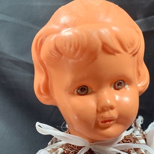 May include: Close-up of a vintage plastic doll's head with brown hair, painted eyes, and a painted smile. The doll is wearing a brown and white dress.