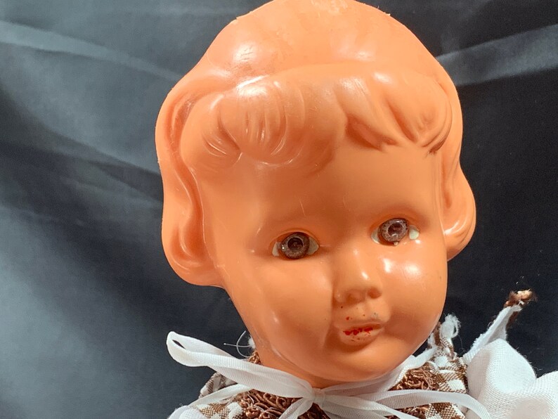 May include: Close-up of a vintage plastic doll's head with brown hair, painted eyes, and a painted smile. The doll is wearing a brown and white dress.