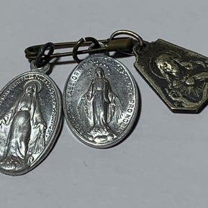 May include: Vintage religious charms and pendants. The charms are attached to a safety pin. Two oval pendants depict a religious figure with text. A third, smaller pendant has a religious figure. The charms are silver and bronze in color.