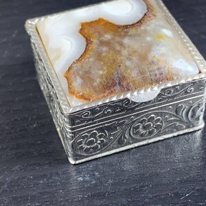 May include: A small, square silver box with a natural stone lid. The lid features a white and brown patterned stone. The sides of the box are decorated with floral and scroll designs. The box is closed.