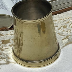Könnte beinhalten: Ein Vintage-Becher aus Messing mit ausgestelltem Körper und rundem Boden. Der Becher hat eine polierte, reflektierende Oberfläche und einen kontrastierenden Silberrand. Der Innenraum ist ein dunkleres, angelaufenes Metall. Der Becher ist wahrscheinlich ein Antiquität oder ein Dekorationsgegenstand.