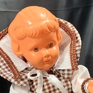 May include: A vintage plastic doll with brown hair, brown eyes, and a brown and white gingham dress. The doll is wearing a white collar with brown trim and a white bow.