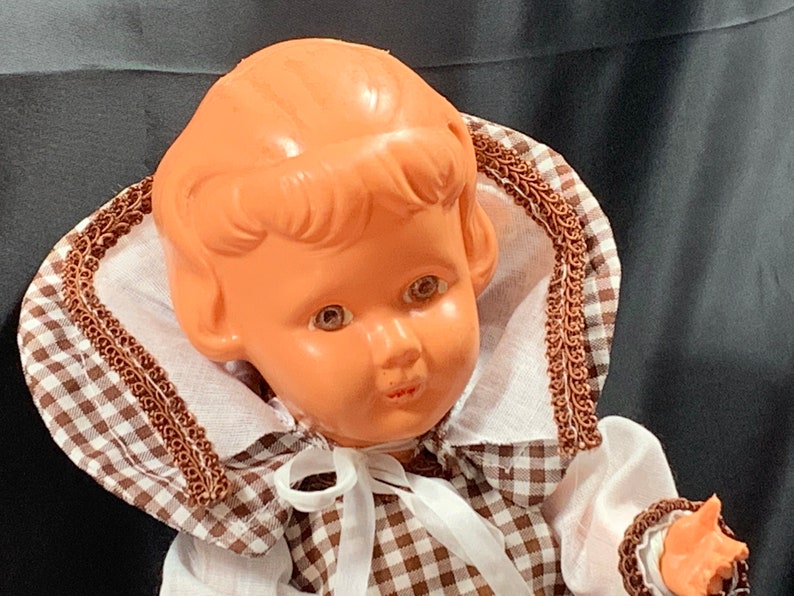 May include: A vintage plastic doll with brown hair, brown eyes, and a brown and white gingham dress. The doll is wearing a white collar with brown trim and a white bow.