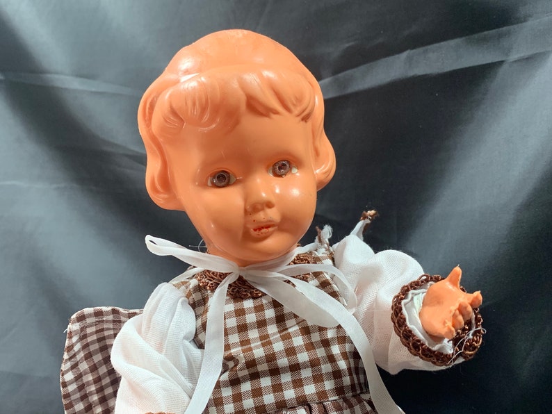 May include: A vintage plastic doll with brown eyes, wearing a white blouse and a brown and white gingham dress.