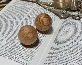 May include: Two wooden, egg-shaped objects with visible wood grain sit on an open book. The objects are light brown and have a smooth, rounded shape. A decorative cord is visible in the background.