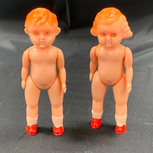 Pode incluir: Duas bonecas de pl&aacute;stico com sapatos vermelhos e cabelo laranja. As bonecas est&atilde;o em p&eacute; sobre uma superf&iacute;cie preta.