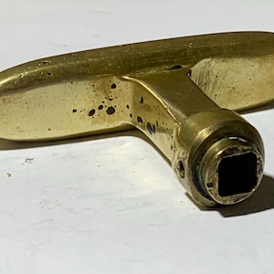 Op de afbeelding: Een T-vormige messing handgreep met een gladde, langwerpige horizontale greep en een cilindrische basis met een vierkante opening. Het metalen oppervlak heeft een gepolijste goudkleurige afwerking met zichtbare donkere vlekken en krasjes.
