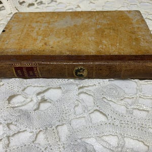 Può includere: Un libro antico con una copertina consumata color marrone chiaro e un dorso in pelle marrone. Il dorso presenta scritte dorate e un piccolo stemma. Il libro è appoggiato su una superficie bianca, simile al pizzo.