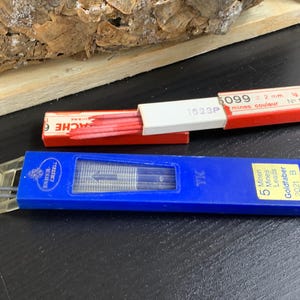 Könnte beinhalten: Ein blaues Plastiketui mit einem transparenten Fenster, in dem fünf schwarze Bleistifte zu sehen sind. Auf dem Etui steht "5 Minen Mines Leads Goldfaber 2021 B". Außerdem ist eine rot-weiße Schachtel mit der Aufschrift "1623P 5099 2 mm. ig 120 mm mines couleur N° 8 ROUGE" zu sehen.