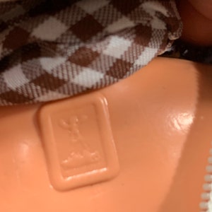 May include: Close-up of a brown and white checkered fabric with a brown plastic tag that has a stylized image of a person standing on a platform. The tag is attached to a brown fabric item.