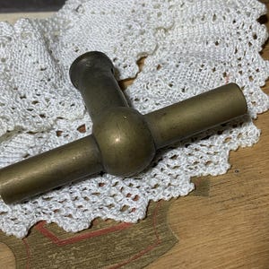 Può includere: Un cavatappi vintage in ottone con un design a forma di T. L'ottone ha un aspetto invecchiato. È appoggiato su un centrino di pizzo bianco, evocando uno stile classico. Il cavatappi è lungo circa 10 cm.