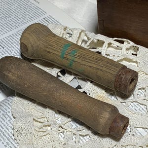 May include: Two antique wooden handles with a cylindrical shape and a rounded end. One handle has faded green markings. The handles are brown and have a rustic, aged appearance. They rest on a lace doily and a book.
