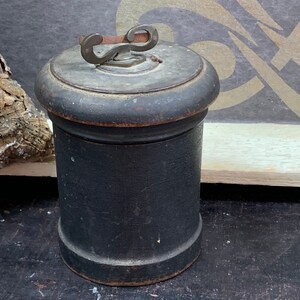 Op de afbeelding: Een zwarte houten container met een metalen deksel en een decoratieve handgreep. De container is cilindrisch en heeft een glad oppervlak.