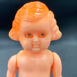 Pode incluir: Uma pequena boneca de pl&aacute;stico com cabelo laranja e olhos pintados. A boneca est&aacute; usando um vestido simples e tem uma express&atilde;o doce.