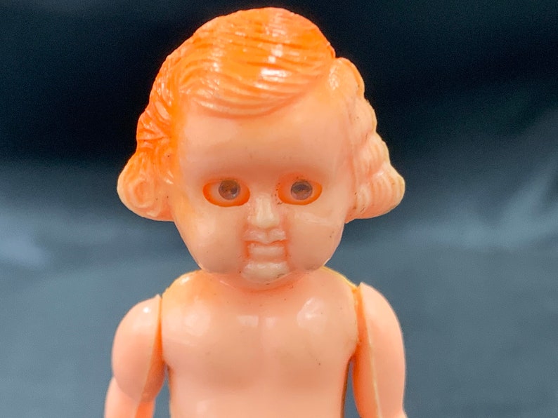 Pode incluir: Uma pequena boneca de pl&aacute;stico com cabelo laranja e olhos pintados. A boneca est&aacute; usando um vestido simples e tem uma express&atilde;o doce.