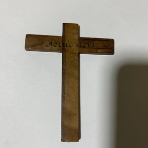 Op de afbeelding: Een houten kruis met het woord "Jerusalem" gegraveerd over de horizontale balk. Het kruis is gemaakt van lichtbruin hout en heeft een eenvoudig, traditioneel ontwerp. Het kruis ligt op een wit oppervlak.