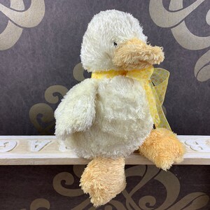 Peut inclure: Un canard en peluche jaune avec un ruban jaune et blanc à pois autour du cou. Le canard est assis sur une étagère blanche avec un motif décoratif.