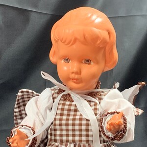 May include: A vintage doll with a brown and white checkered dress, white lace trim, and a white ribbon around the neck. The doll has a light brown plastic head with painted brown hair, blue eyes, and a pink complexion.