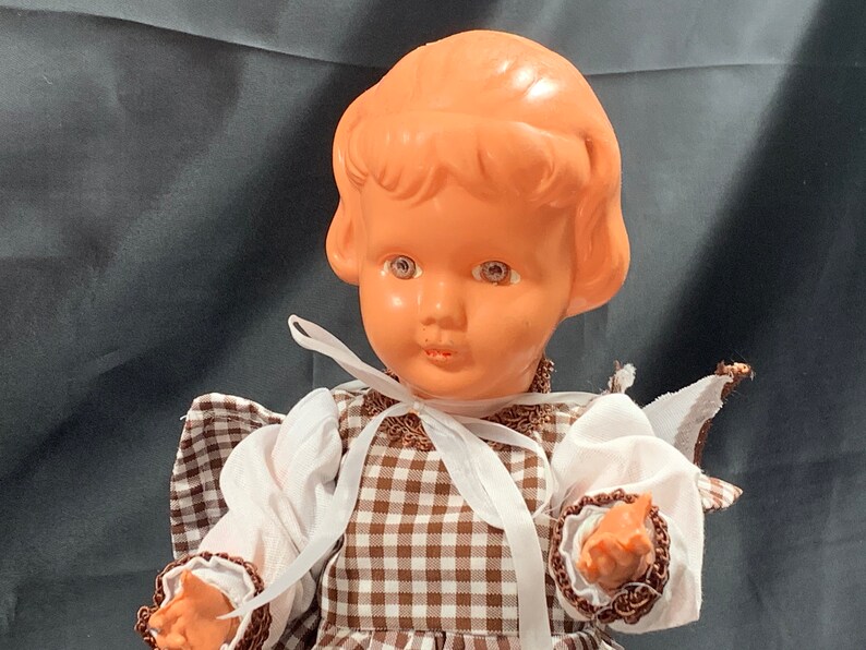 May include: A vintage doll with a brown and white checkered dress, white lace trim, and a white ribbon around the neck. The doll has a light brown plastic head with painted brown hair, blue eyes, and a pink complexion.