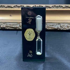 May include: A black rectangular stand with a gold clock face, a silver hourglass, and gold gears. The clock face reads "G.J. Geneva"