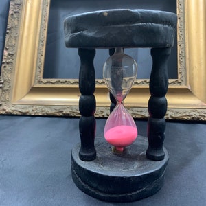 May include: A black wooden hourglass with pink sand. The hourglass is on a black surface.