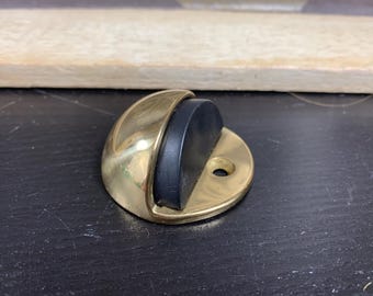 May include: A brass door stop with a black rubber bumper. The doorstop has a half-moon shape with a circular base and a hole for mounting. The brass has a polished finish, and the rubber is matte black.