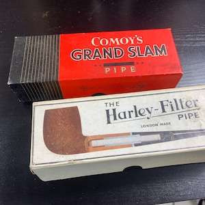 Pode incluir: Duas caixas de cachimbo vintage. A caixa superior é vermelha com "COMOY'S GRAND SLAM PIPE" impresso em branco. A caixa inferior é branca com "THE Harley-Filter PIPE" impresso em preto, com uma ilustração de cachimbo. Ambas as caixas são retangulares.